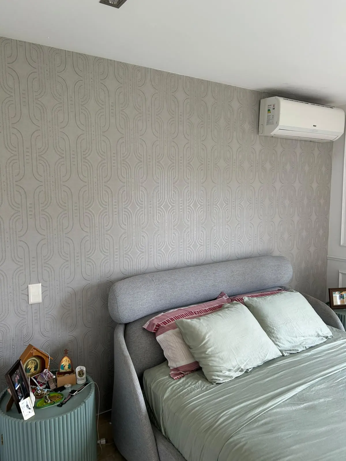 Papel tapiz de trama geométrica en gris y plata cubriendo la pared de un dormitorio, detrás de una cabecera acolchada gris